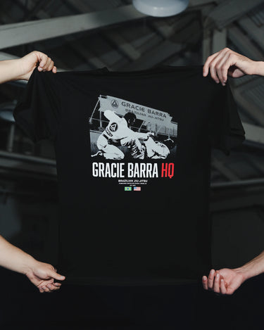 Black GB HQ 20th Anniversary Tee held up by two people in a training-facility setting, showing the full back graphic of GB athletes sparring in front of the original Irvine Headquarters.