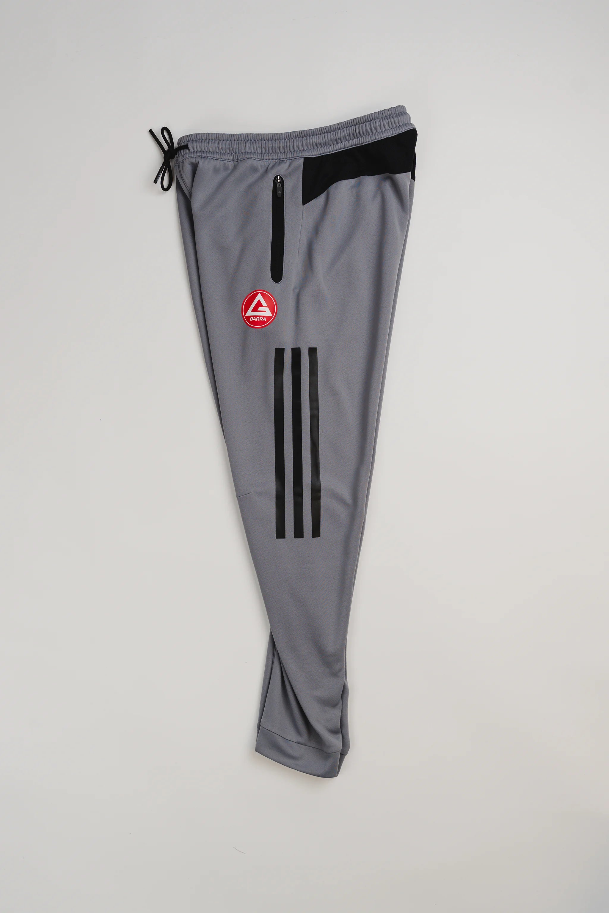 Gray GB Team Track Jogger by adidas folded with black stripes and red Gracie Barra logo visible, light gray background.