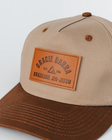 Close-up of the GB Trademark Cap showing the embossed leather Gracie Barra patch on the beige crown and brown curved visor, photographed on a white background.