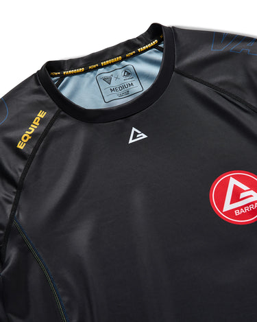 Close-up of black Gracie Barra x Vanguard Evolution rashguard showing reinforced crew neckline, yellow “Equipe” detail, Vanguard collar taping, and red Barra Shield chest logo.