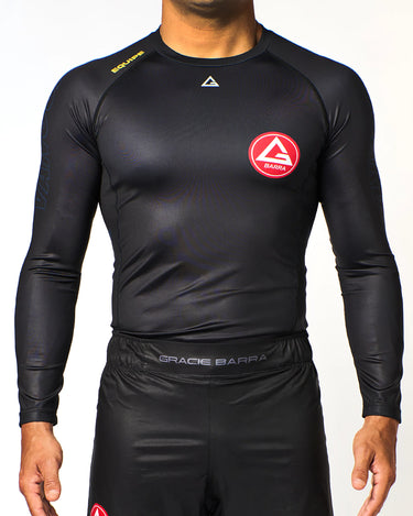 Athlete wearing black Gracie Barra x Vanguard Evolution rashguard with red Barra Shield chest logo, yellow “Equipe” shoulder detail, and matching GB shorts.