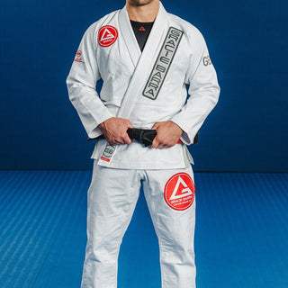 Adult athlete wearing the white GB1 Kimono standing on a blue mat, featuring the Red Shield patch on the chest, Gracie Barra lapel patch, and reinforced jacket and pant design