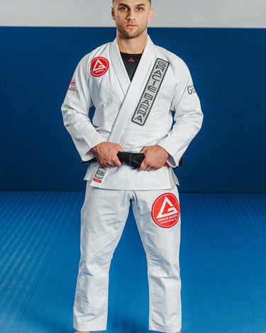 Adult athlete wearing the white GB1 Kimono standing on a blue mat, featuring the Red Shield patch on the chest, Gracie Barra lapel patch, and reinforced jacket and pant design