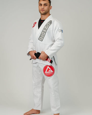 Adult athlete wearing the white GB1 Kimono in a left-facing stance, featuring the Red Shield chest patch, Gracie Barra lapel patch, GB arm logo, and tailored, reinforced construction.