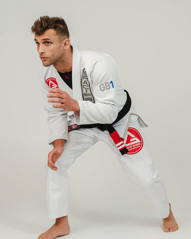 Adult athlete wearing the white GB1 Kimono in a low training stance, showing the Red Shield chest and pant patches, GB1 arm embroidery, and the reinforced, tapered fit built for performance