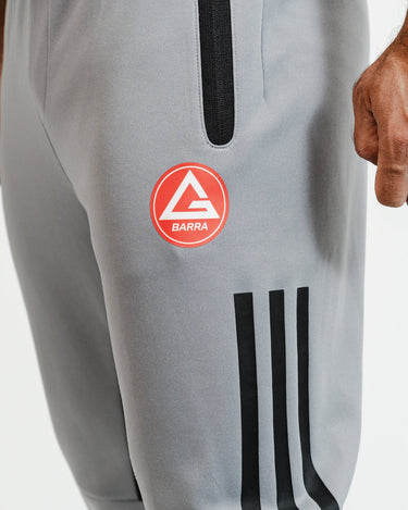 Close-up of red Gracie Barra logo and black stripes on gray GB Team Track Jogger by adidas.