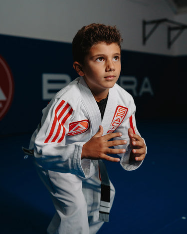 Youth student in white GB x adidas III Stripes Kimono with red 3-Stripes and Barra Shield patch, posed in grappling stance on blue mats inside a Gracie Barra academy.