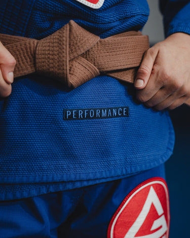 Close-up of blue GB x adidas III Stripes Women’s Kimono during training, showing embroidered Performance text and large Red Shield patch below brown belt.