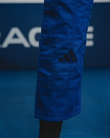 Close-up of blue GB x adidas III Stripes Women’s Kimono pant leg showing embroidered black adidas logo above the cuff during training on blue mats.