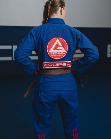 Back view of female athlete wearing blue GB x adidas III Stripes Women’s Kimono on blue mats, featuring large Red Shield patch, red EQUIPE GB patch, and red 3-Stripes on pant legs.