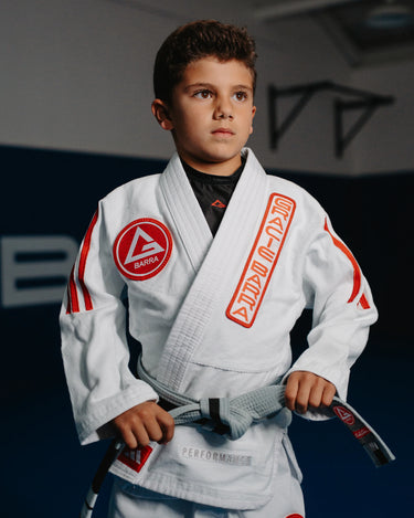 Youth student wearing white GB x adidas III Stripes Kimono with red 3-Stripes and Barra Shield patch, standing on the mats inside a Gracie Barra academy.