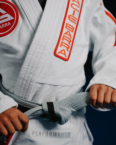 Close-up of youth student tying belt while wearing white GB x adidas III Stripes Kimono with red Gracie Barra lapel patch, Barra Shield on chest, and embroidered Performance mark.