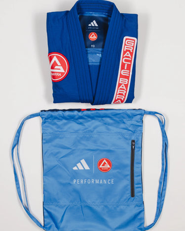 Folded blue GB x adidas III Stripes Youth Kimono with red Gracie Barra patch and light blue drawstring bag featuring adidas and Gracie Barra logos with Performance text on white background.