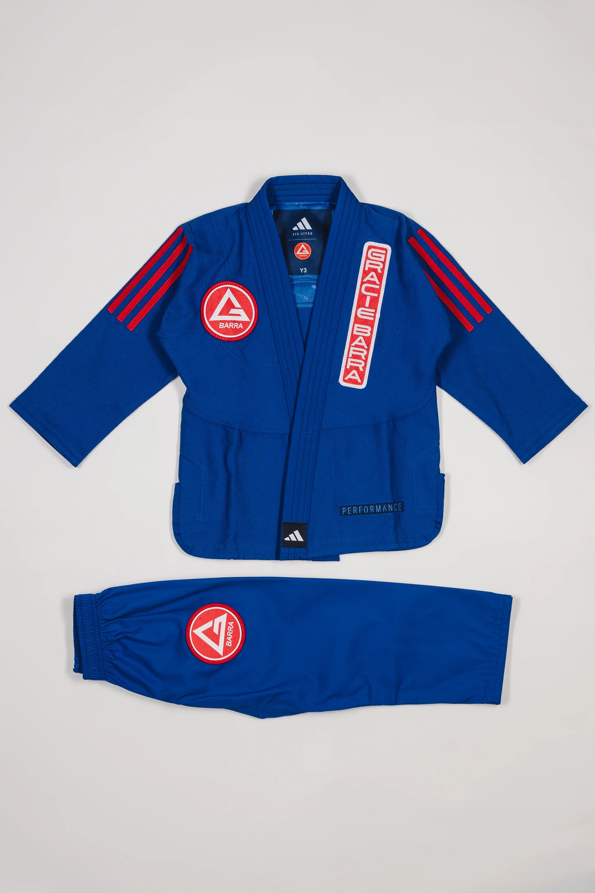 Close-up of blue GB x adidas III Stripes Youth Kimono jacket showing red Gracie Barra patch on lapel, GB Red Shield on chest, and adidas logo at collar on white background.