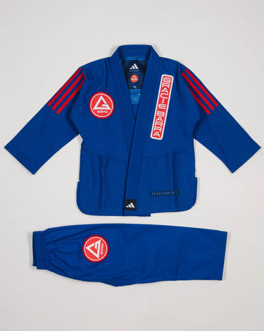 Close-up of blue GB x adidas III Stripes Youth Kimono jacket showing red Gracie Barra patch on lapel, GB Red Shield on chest, and adidas logo at collar on white background.