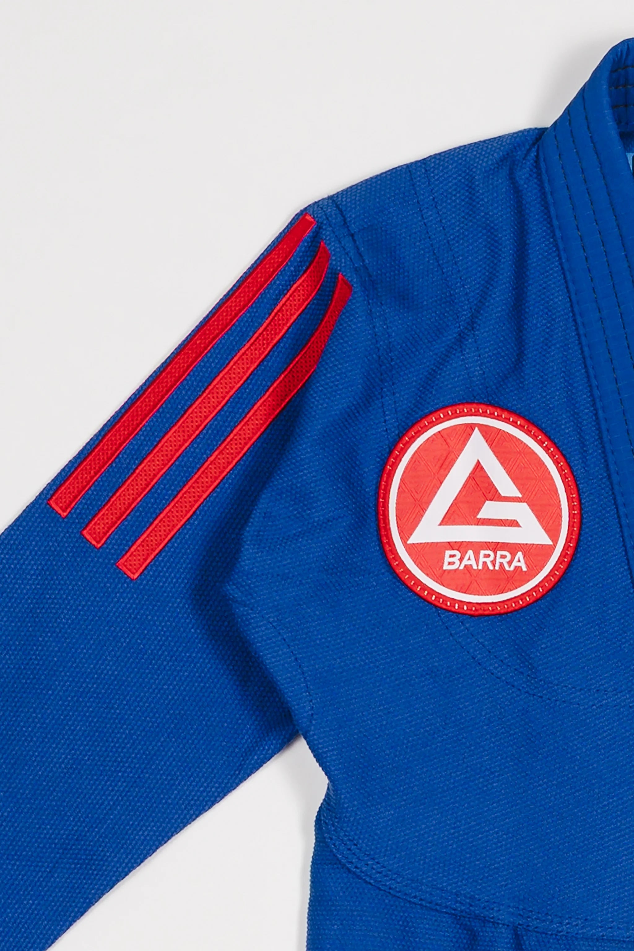 Close-up of blue GB x adidas III Stripes Youth Kimono shoulder showing bold red 3-Stripes detail and Gracie Barra Red Shield patch on white background.