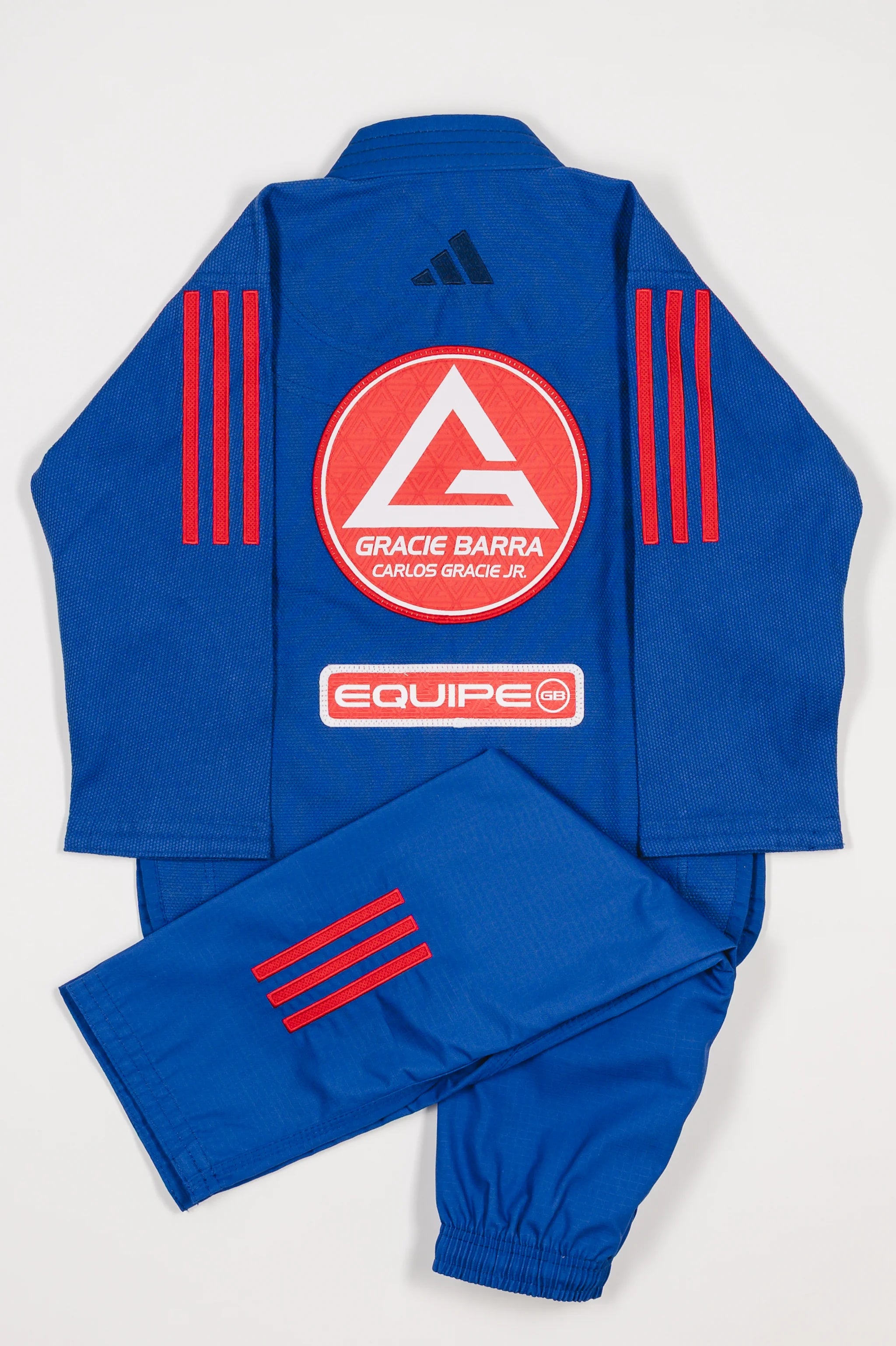 Back view of blue GB x adidas III Stripes Youth Kimono with pants folded across jacket, featuring large Red Shield patch, red 3-Stripes on shoulders and pant leg, and adidas logo at upper back.