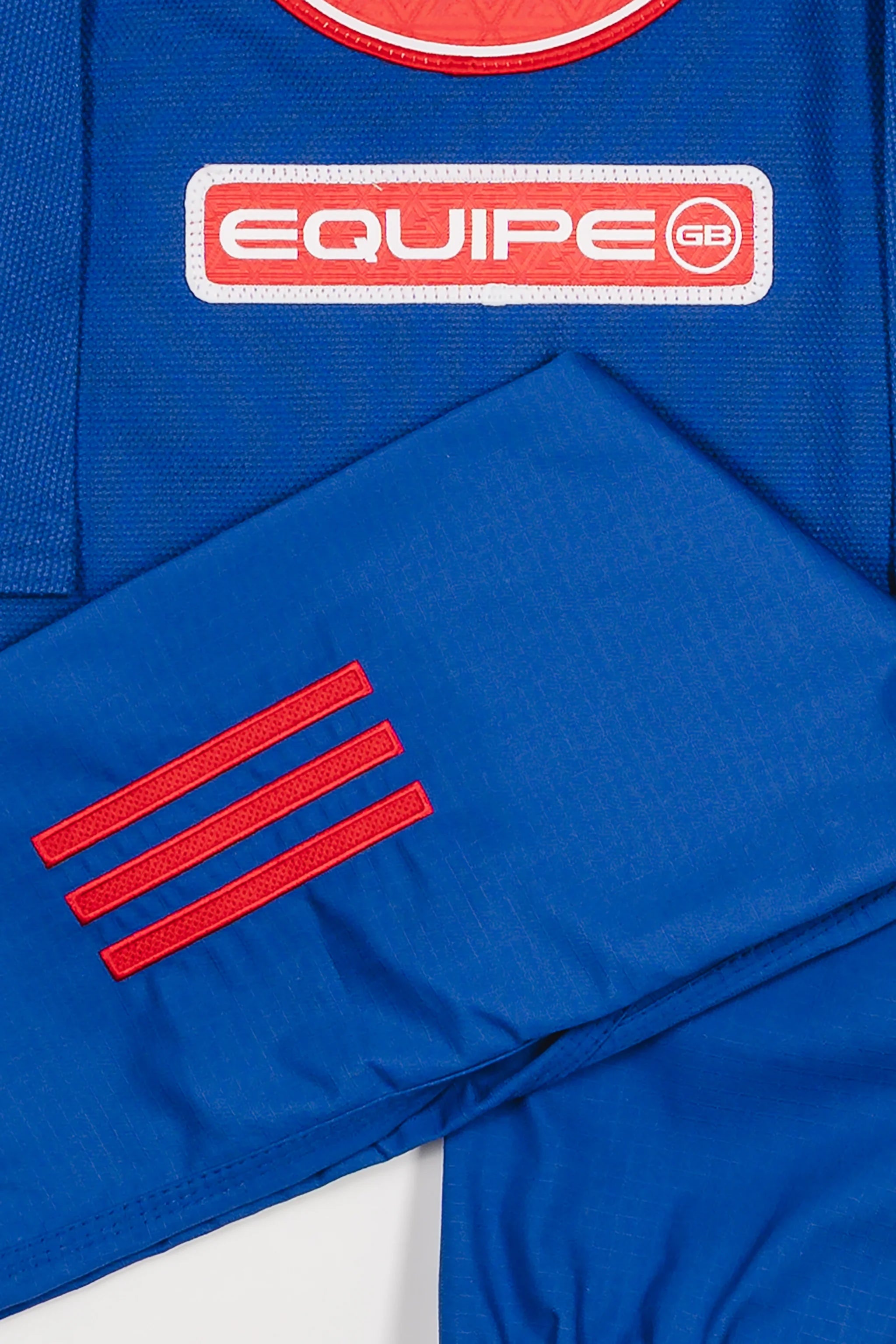 Close-up of blue GB x adidas III Stripes Youth Kimono pants and jacket showing red 3-Stripes detail on pant leg and red EQUIPE GB patch on jacket back.