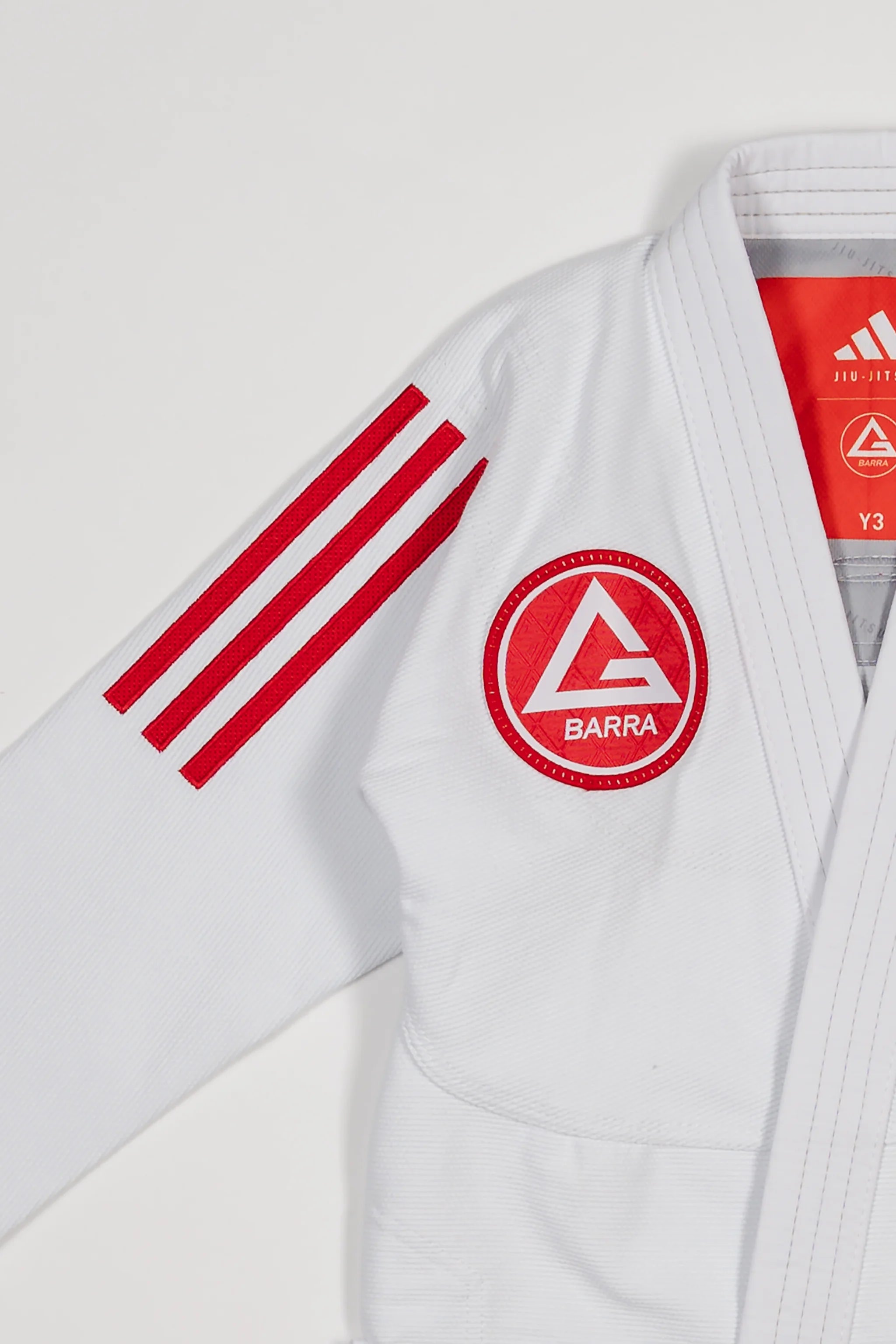Close-up of white GB x adidas III Stripes Youth Kimono shoulder showing bold red 3-Stripes and Barra Shield patch on chest with adidas Jiu-Jitsu label visible inside collar.
