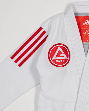 Close-up of white GB x adidas III Stripes Youth Kimono shoulder showing bold red 3-Stripes and Barra Shield patch on chest with adidas Jiu-Jitsu label visible inside collar.