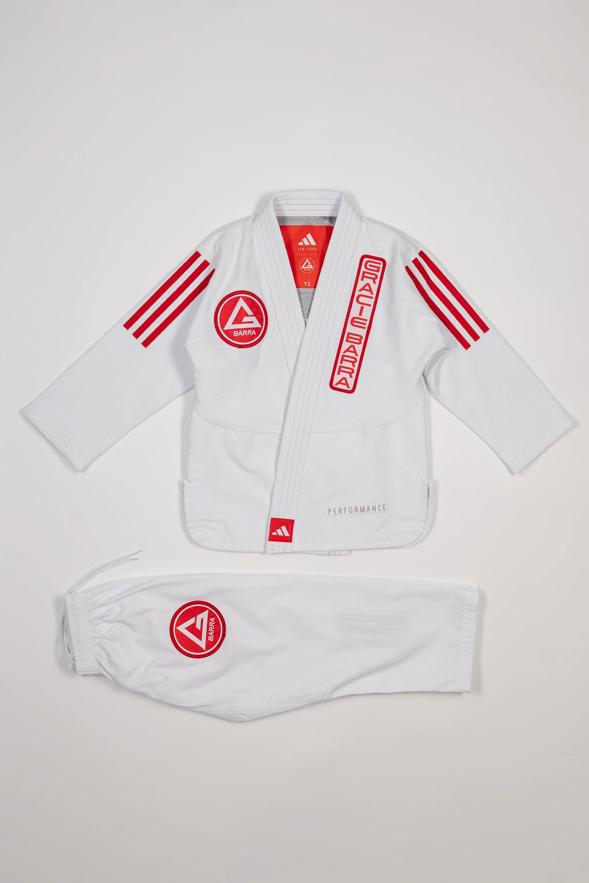White GB x adidas III Stripes Youth Kimono laid flat on white background, featuring bold red 3-Stripes on shoulders and red Gracie Barra patches on jacket and pants.