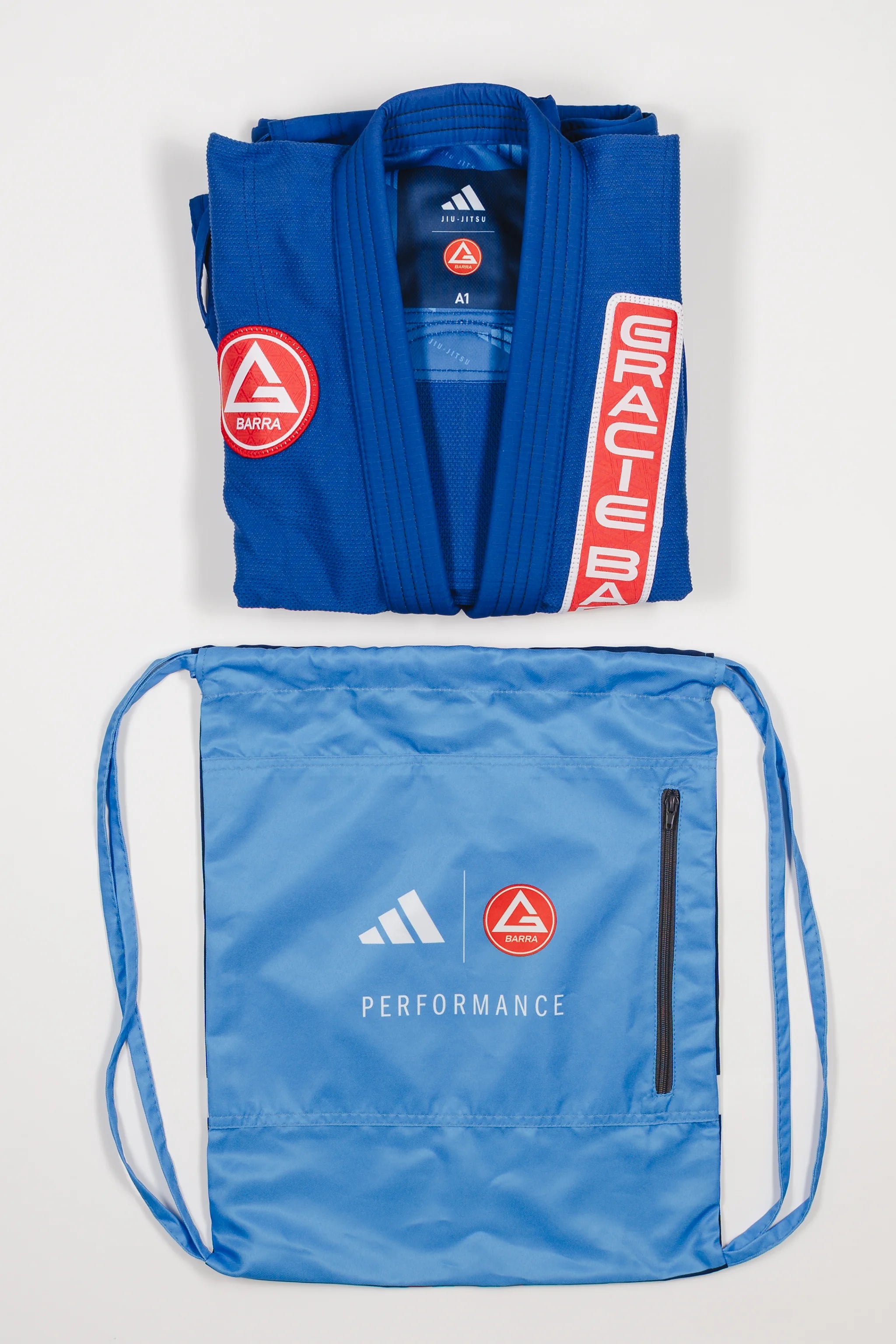 Folded blue GB x adidas III Stripes Women’s Kimono with red Barra Shield patch and light blue drawstring bag featuring adidas and Gracie Barra logos with Performance text on white background.