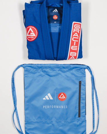 Folded blue GB x adidas III Stripes Women’s Kimono with red Barra Shield patch and light blue drawstring bag featuring adidas and Gracie Barra logos with Performance text on white background.