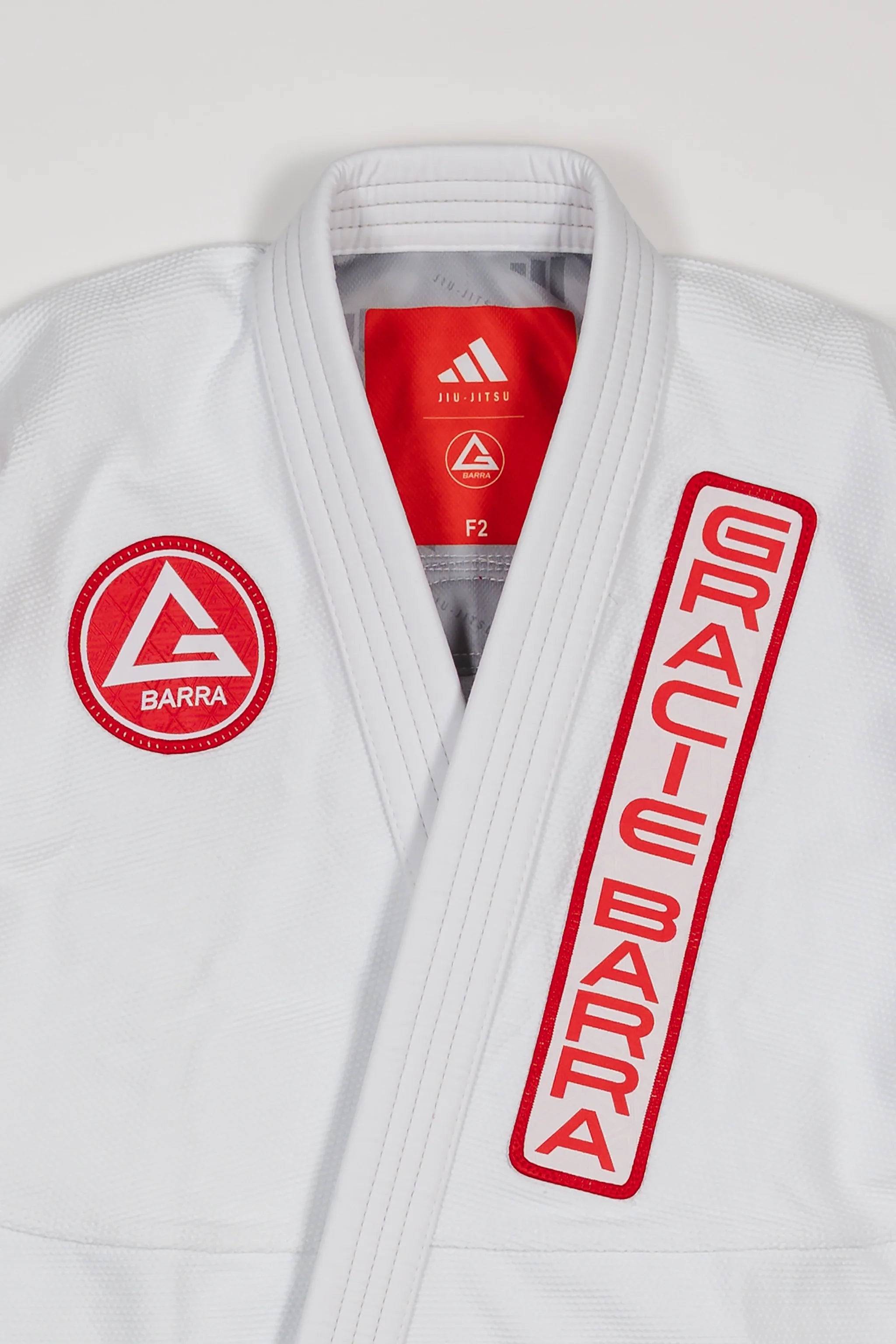 Close-up of white GB x adidas III Stripes Women’s Kimono jacket showing red Barra Shield on chest, Gracie Barra lapel patch, and adidas Jiu-Jitsu label inside collar.