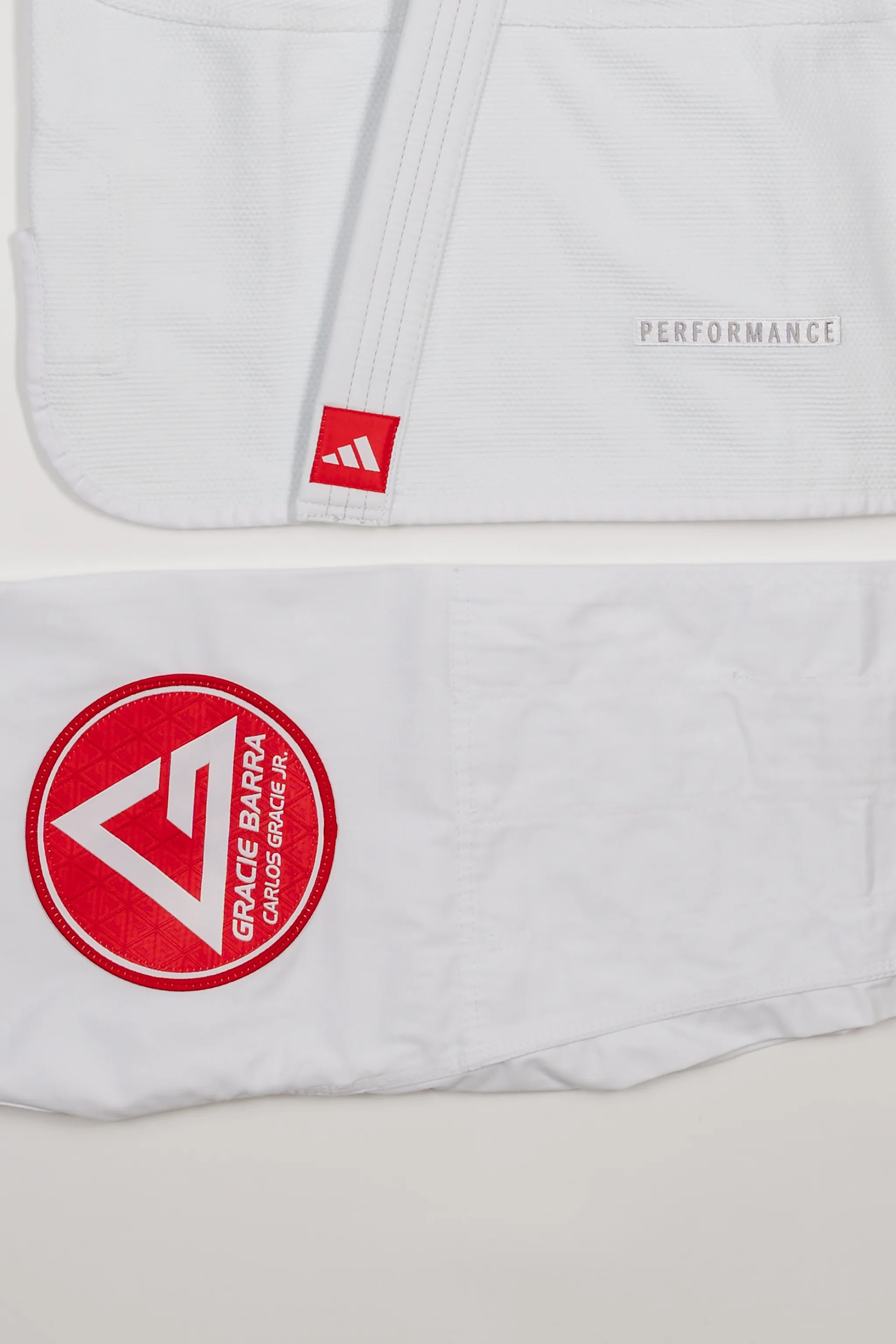 Detail view of white GB x adidas III Stripes Women’s Kimono jacket and pants on white background, showing embroidered adidas Performance mark on jacket hem and large Red Shield patch on pant leg.