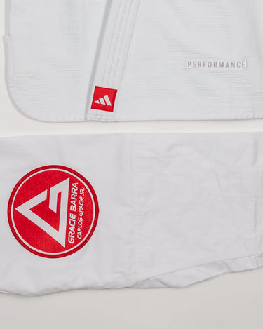 Detail view of white GB x adidas III Stripes Women’s Kimono jacket and pants on white background, showing embroidered adidas Performance mark on jacket hem and large Red Shield patch on pant leg.