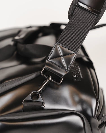 Close-up of the Legacy Travel Duffel’s padded shoulder strap attachment, showing reinforced stitching, metal hardware, and the smooth black exterior material.