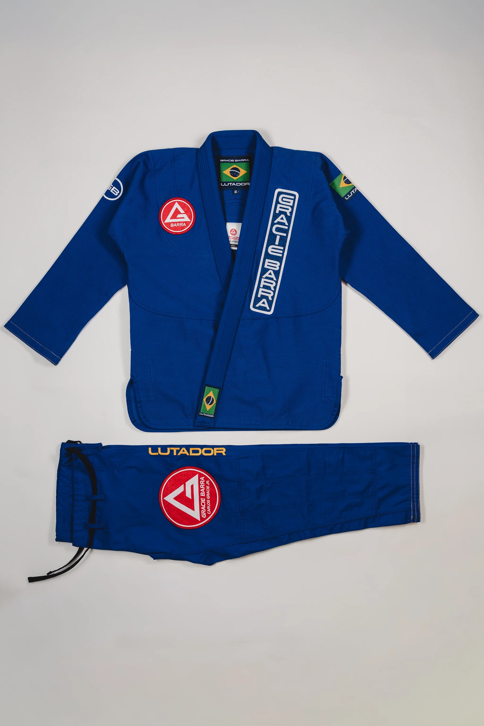Blue Lutador Kimono set laid flat on a light gray background, featuring the pearl-weave jacket with Gracie Barra patches and embroidery, and matching ripstop pants with the Lutador wordmark and red Barra Shield.