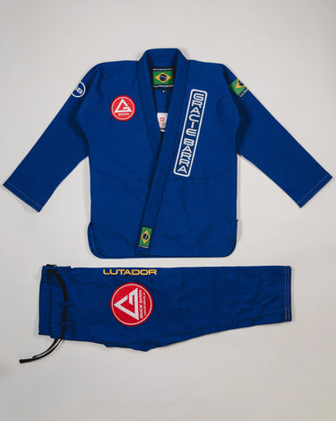 Blue Lutador Kimono set laid flat on a light gray background, featuring the pearl-weave jacket with Gracie Barra patches and embroidery, and matching ripstop pants with the Lutador wordmark and red Barra Shield.