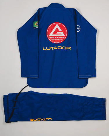 Back view of the blue Lutador Kimono jacket and pants laid flat on a light gray background, featuring the large red Barra Shield, gold Lutador embroidery, and matching pant branding.