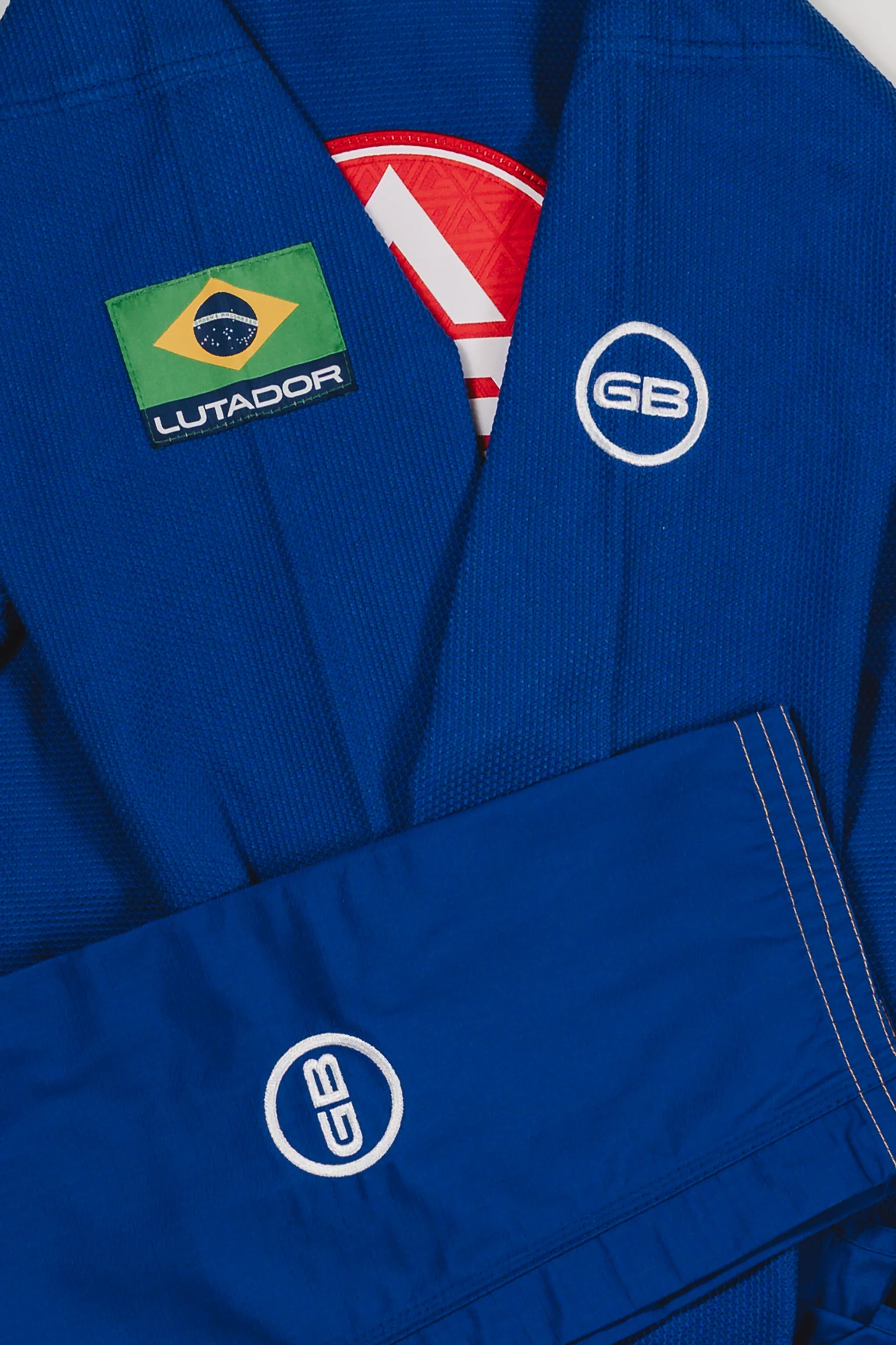 Close-up of the blue Lutador Kimono showing the Brazilian flag Lutador patch, white GB circle logos on the sleeve and pant leg, and textured pearl-weave fabric.