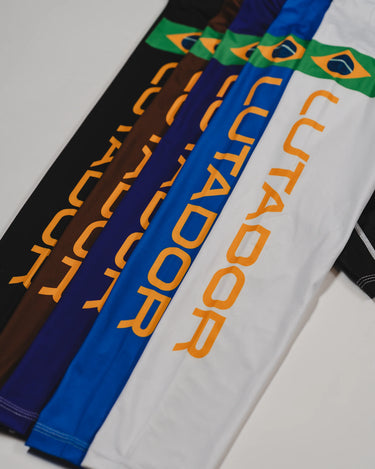 Angled layout of the Lutador Longsleeve Ranked Rashguard sleeves in multiple belt-ranked colorways—white, blue, purple, brown, and black—each featuring the gold ‘Lutador’ print and Brazilian flag patch.