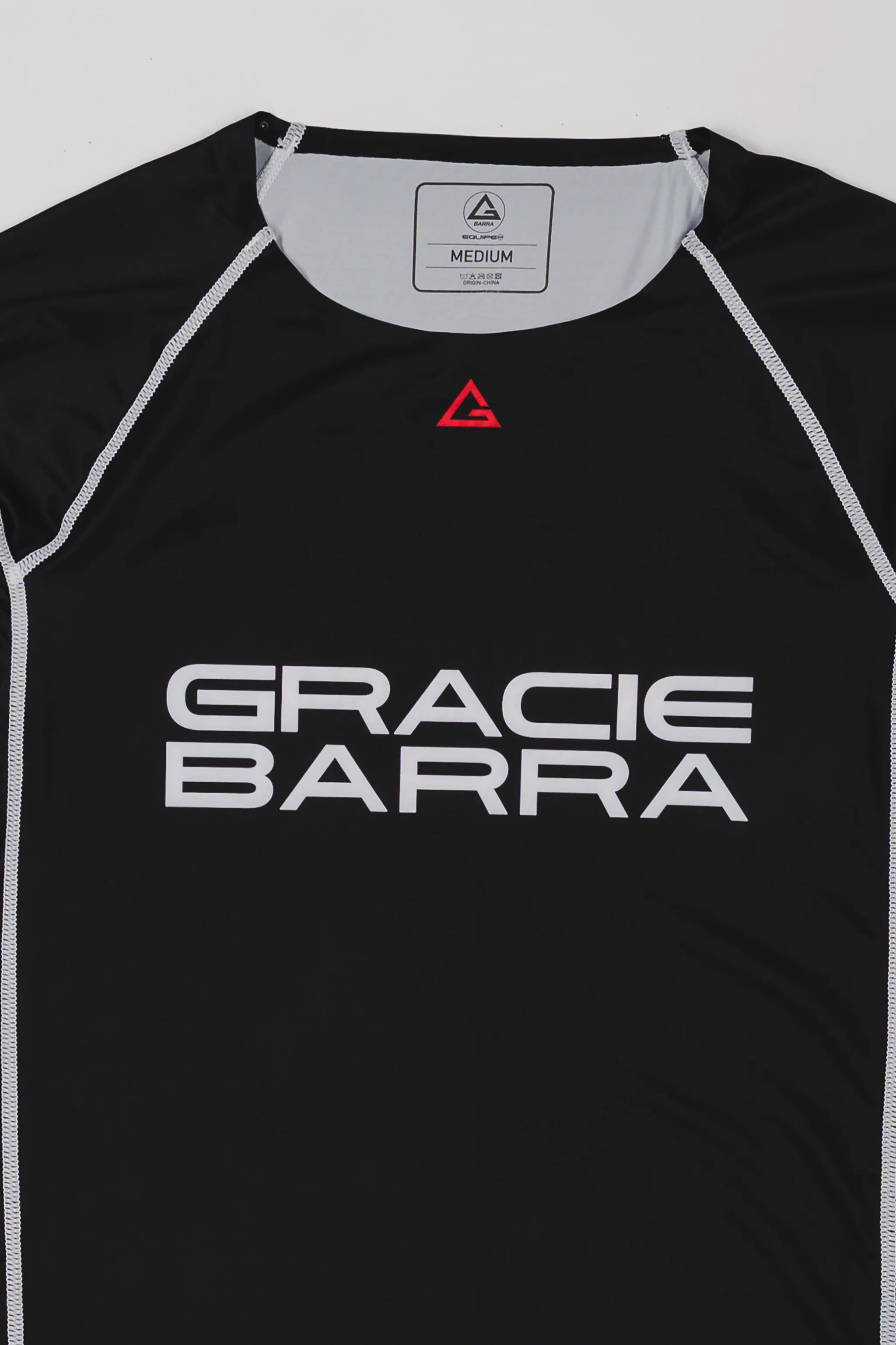 Close-up of the black Lutador long-sleeve ranked rashguard showing the GRACIE BARRA chest print, red Legacy G triangle emblem at the collar, and white contrast stitching.