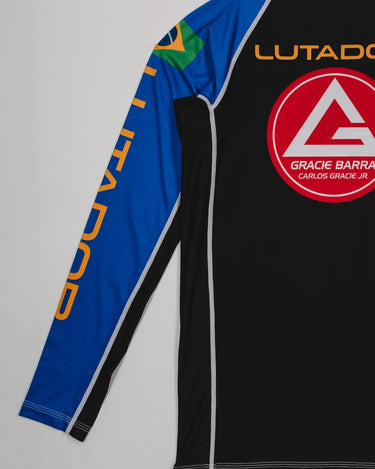 Angled back view of the Lutador Longsleeve Ranked Rashguard in blue, highlighting the gold ‘Lutador’ sleeve print, Brazilian flag arm patch, and large red Barra Shield on the black panel.