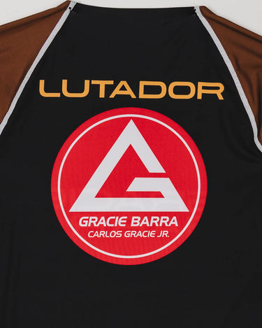 Close-up of the back of the Lutador Longsleeve Ranked Rashguard in brown, showing the gold ‘Lutador’ text and large red Barra Shield on the black panel with brown sleeves.