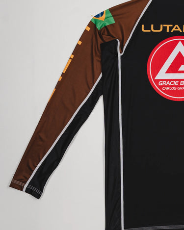 Angled back view of the Lutador Longsleeve Ranked Rashguard in brown, showing the gold ‘Lutador’ sleeve print, Brazilian flag patch, and large red Barra Shield on the black torso.