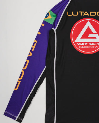 Angled back view of the Lutador Longsleeve Ranked Rashguard in purple, showing the gold ‘Lutador’ sleeve print, Brazilian flag patch, and large red Barra Shield on the black torso.
