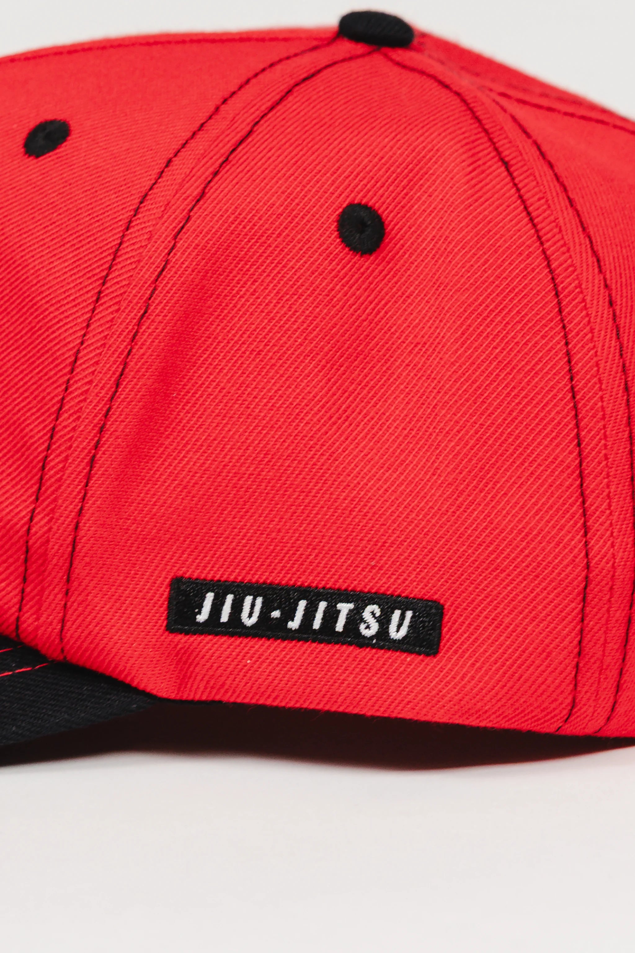 Close-up of the Red Comp Team Cap showing the woven black JIU-JITSU label stitched onto the GB Red wool twill side panel with black eyelets and stitching.