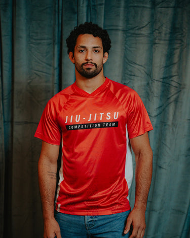 Professor Ian wearing the Men’s Red Comp Team Jersey featuring the official Jiu-Jitsu Competition Team graphic, standing against a dark curtain backdrop.