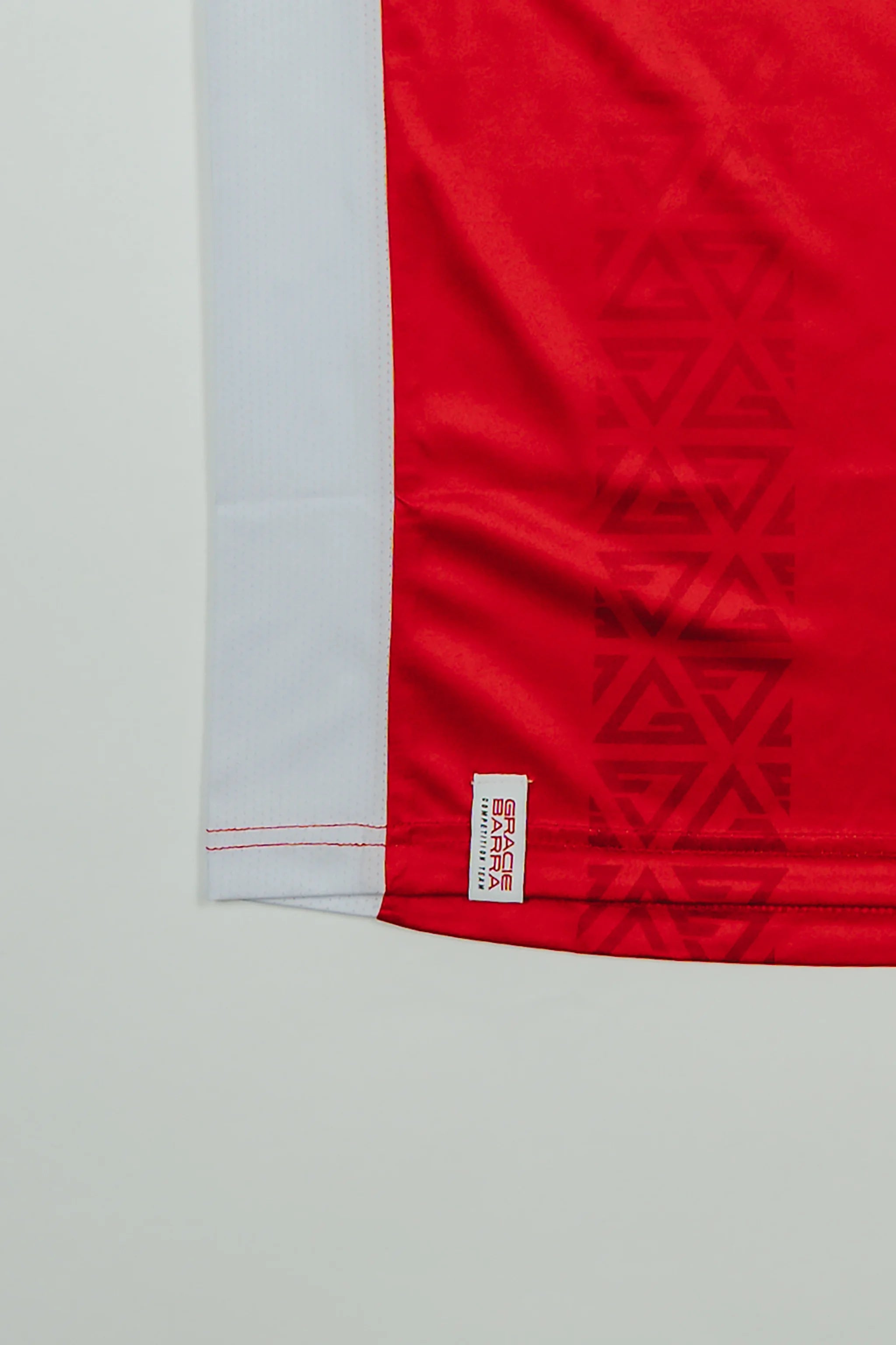 Close-up of the Men’s Red Comp Team Jersey hem showing the tonal Legacy G geometric pattern, white mesh side panel, and woven Gracie Barra Competition Team label.