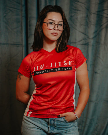 Professor Keanna wearing the Red Comp Women’s Team Jersey featuring the official Jiu-Jitsu Competition Team graphic, photographed against a dark backdrop.