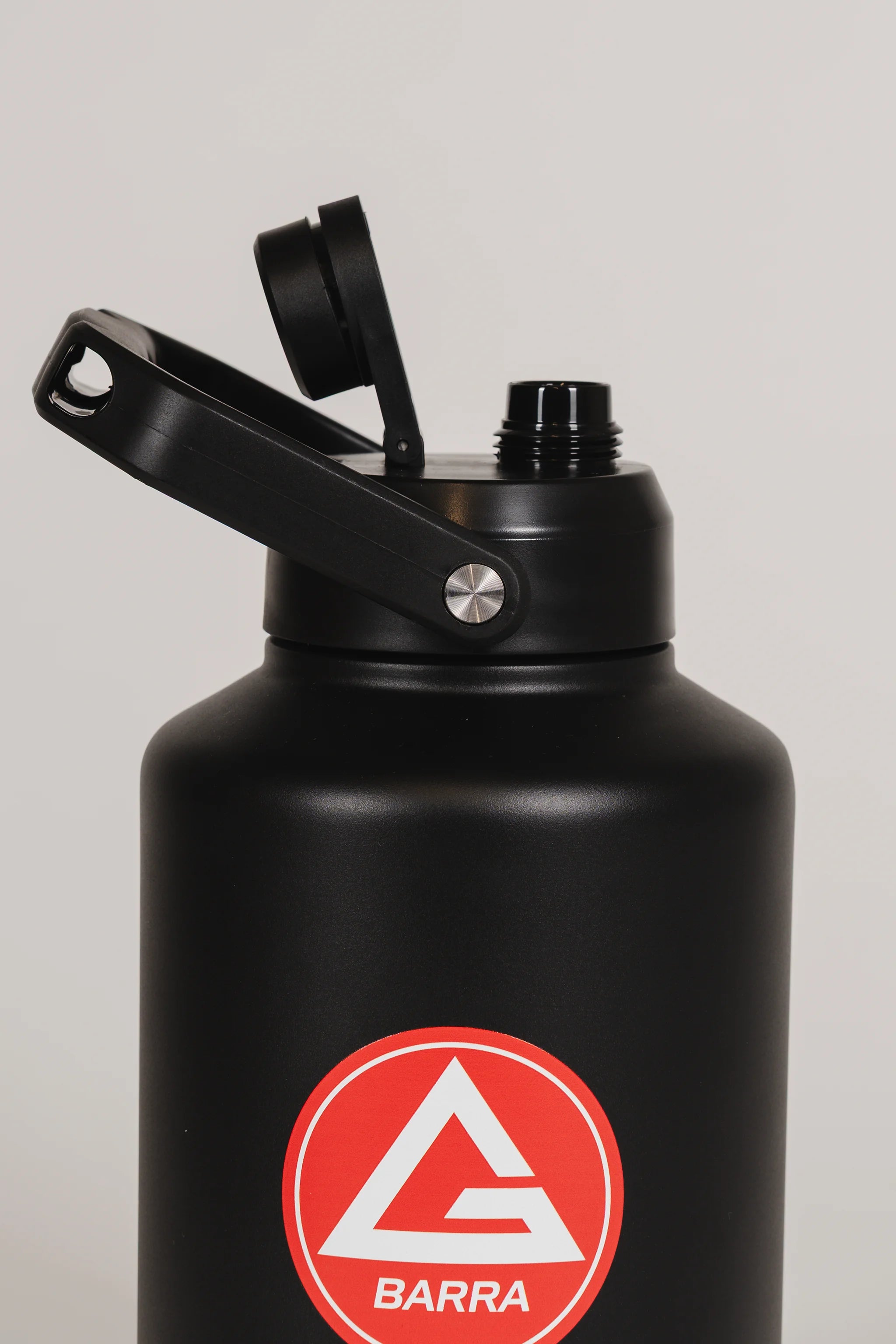 Close-up of the black stainless steel 128 oz Red Shield bottle with the lid open and the Gracie Barra Red Shield logo visible on the front, photographed against a light background.