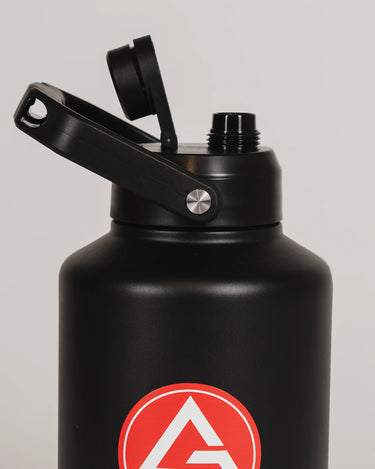 Close-up of the black stainless steel 128 oz Red Shield bottle with the lid open and the Gracie Barra Red Shield logo visible on the front, photographed against a light background.