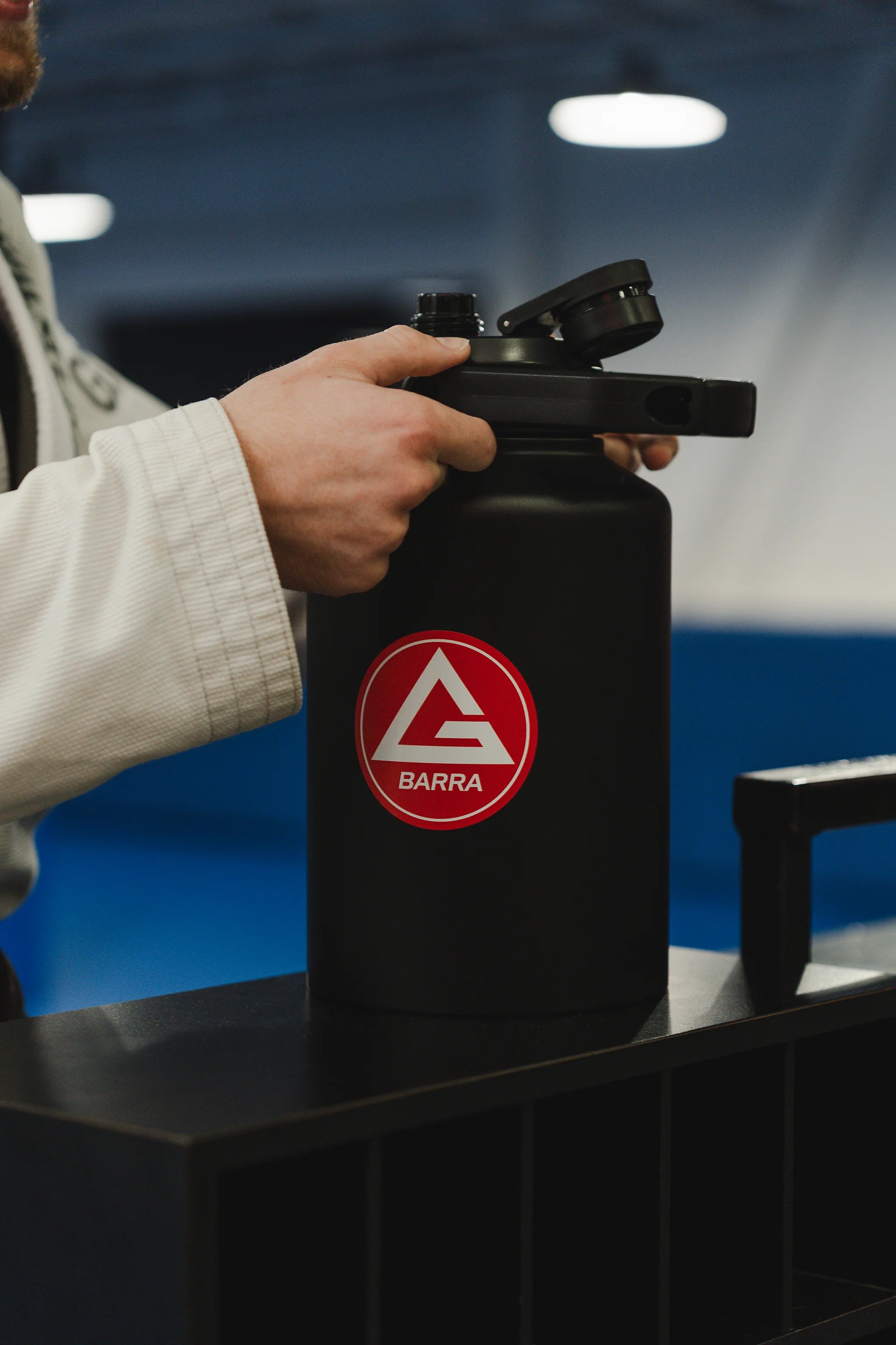 Professor in a white Jiu-Jitsu gi holding the black 128 oz Red Shield bottle with the Gracie Barra Red Shield logo, photographed inside a GB gym.