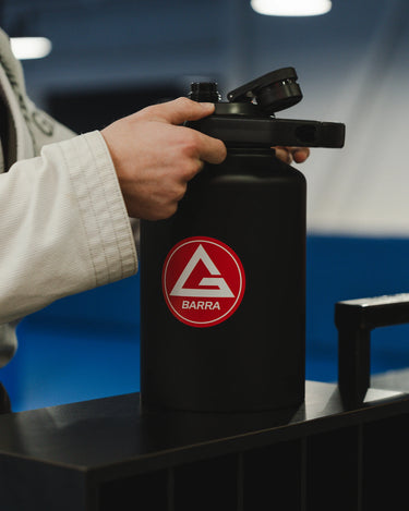 Professor in a white Jiu-Jitsu gi holding the black 128 oz Red Shield bottle with the Gracie Barra Red Shield logo, photographed inside a GB gym.