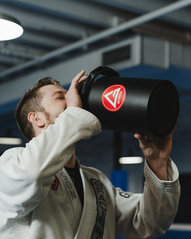 Professor in a white Jiu-Jitsu gi drinking from the black 128 oz Red Shield bottle with the Gracie Barra Red Shield logo, inside a GB training academy.